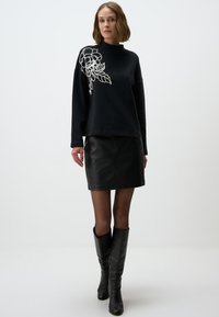 Maglione nero con ricami floreali bianchi sulla spalla sinistra, abbinato a una mini gonna in pelle nera e stivali neri alti fino al ginocchio.
