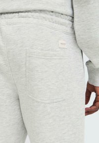 Tejido de sudadera gris que presenta un bolsillo trasero visible con una etiqueta de logo cuadrada. La textura es suave y ligeramente jaspeada.