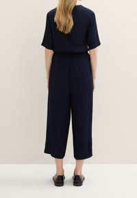 Námořnický overal s krátkým rukávem, volným střihem, širokými zkrácenými nohavicemi, strukturovanou tkaninou a stáhnutým pasem. Černé boty doplňují outfit.