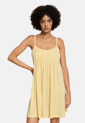 Vrouw met krullend haar, gekleed in een mouwloze, loszittende gele jurk met plooien bij de borst, staand tegen een effen achtergrond.