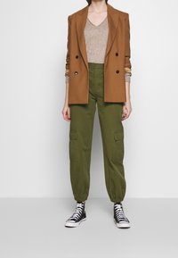 Brun dubbelknäppt kavaj, beige v-ringad tröja, olivgröna cargobyxor med elastiska fållar, bärs med svarta hög-top sneakers.