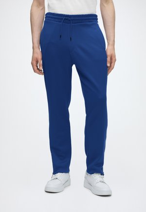 Persoană purtând pantaloni de trening albastru regal cu șnur, adidași albi și o cămașă albă, stând pe un fundal deschis simplu.