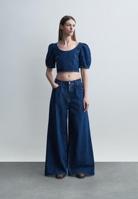 Moteris dėvi tamsiai mėlyną džinso trumpą palaidinę su pūstomis rankovėmis ir platėjusias aukšto liemens džinsines kelnes, stovinti prie paprasto pilko fono.