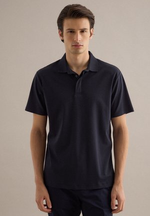 Joven de pie, vistiendo una camisa tipo polo de manga corta en color azul marino oscuro lisa, sobre un fondo neutro.
