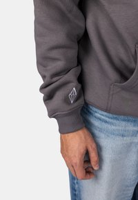 Felpa grigia con polsini a costine. Presenta un piccolo logo geometrico bianco sulla manica. Indossata con jeans in denim blu chiaro.