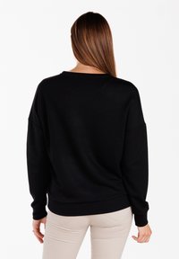 Sudadera negra de corte relajado, con escote redondo y hombros caídos. Textura suave y dobladillo acanalado, combinada con pantalones de color beige claro.