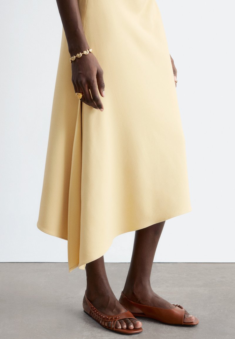 Femme en jupe asymétrique jaune pâle, portant des ballerines ouvertes marron, un bracelet en or et une grosse bague en or sur une peau foncée.