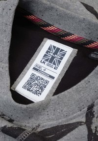 Grijze sweatshijfstof met een zwarte binnenlaag, voorzien van een patroonnek met kleurrijke accenten en een label met merk en QR-code.