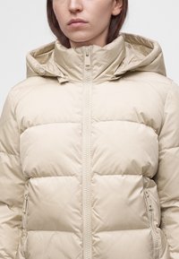 Femme portant une doudoune beige clair avec un col haut, une capuche attachée et une fermeture éclair à l'avant et aux poches.