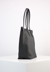 Sac fourre-tout noir à surface texturée, avec des anses lisses, une forme allongée et une fermeture éclair. Présenté seul sur un socle blanc.
