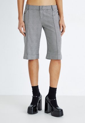 Person iført grå ternede knælange shorts, sorte sokker og sorte lakplatformhæle mod en hvid baggrund.