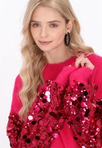 Fukszia pulóver bolyhos textúrával és flitteres rózsaszín díszítéssel az ujjakon. A modellnek hullámos szőke haja van, és nagy fekete fülbevalót visel.