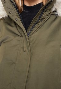Parka verde oliva con zip frontale, cappuccio con bordo in pelliccia e colletto nero a costine. Presenta bottoni a pressione e una texture liscia.