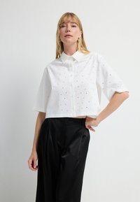 Camisa blanca cortada con cuello puntiagudo y mangas cortas, adornada con pequeños detalles bordados en negro y plata. Combinada con pantalones negros.