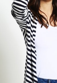 Mujer vestida con una camisa blanca y un cárdigan de rayas navy y blanco de manga larga, con el brazo levantado y el cabello castaño visible.