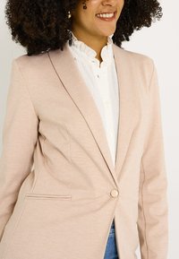 Veste rose pâle avec une coupe ajustée, fermeture à un seul bouton et revers crantés. Elle présente une texture subtile et un détail de poche passepoilée.