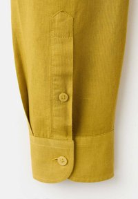 Senfgelber Leinenhemdärmel mit Knopfverschluss, spitz zulaufender und abgerundeter Form, der strukturierte Stoff und Nähte sind deutlich sichtbar.