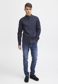 Camisa de botones azul marino con cuello band, combinada con jeans ajustados azules y zapatos negros. La camisa tiene una textura suave y un diseño ajustado.