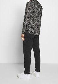 Černá vzorovaná košile s dlouhým rukávem a květinovými motivy, v kombinaci s černými kalhotami a bílými teniskami. Materiál je hladký na dotek.