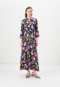 Woman wearing a long, floral dark blue dress with pink, blue, and yellow flowers, standing against a plain white background.