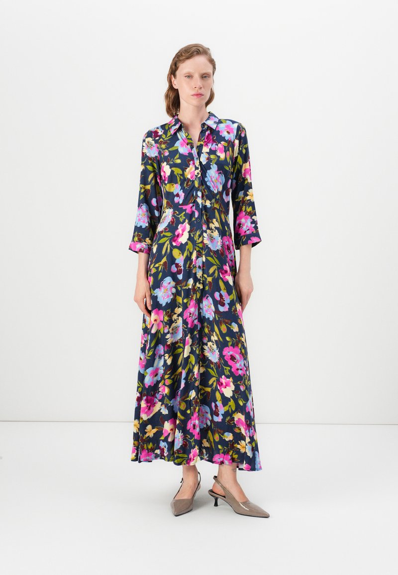 Woman wearing a long, floral dark blue dress with pink, blue, and yellow flowers, standing against a plain white background.
