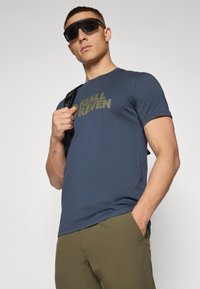 Námornícky modré tričko Fjällräven s logom v olivovo zelenej farbe, krátke rukávy, okrúhly výstrih, kombinované s olivovo zelenými nohavicami a čiernymi slnečnými okuliarmi.