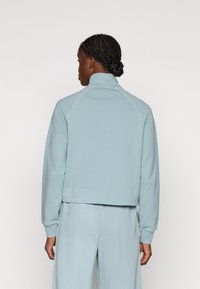 Lys blå zippered sweatshirt med høj krave, der har lange ærmer og hvide syninger, lavet af blødt, tekstureret stof.