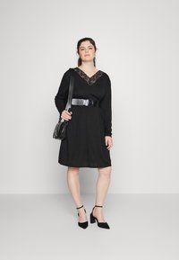 Vestido preto de mangas longas com decote V em renda, ajustado na cintura com um cinto largo preto. Combinado com sapatos de salto agudo pretos e uma bolsa de ombro.