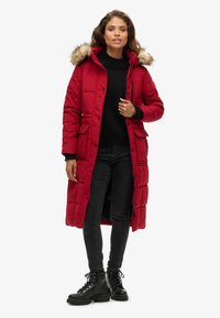 Abrigo acolchado rojo con capucha forrada de piel, que presenta cremalleras robustas, botones de presión y dos bolsillos frontales. Combinado con jeans negros y botas.