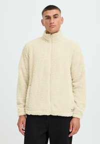 Giacca in pile beige chiaro con colletto alto e zip completa. Presenta un esterno testurizzato e morbido e polsini elastici.