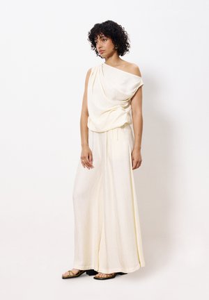 Une tenue texturée de couleur crème avec un top asymétrique à une épaule et un pantalon large. Le modèle porte des sandales plates. Un design simple et élégant.