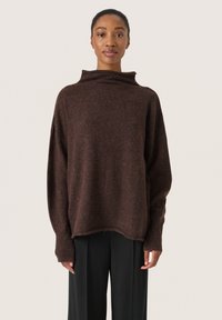 Brauner Strickpullover mit hohem Kragen und überschnittenen Schultern. Verfügt über eine weiche Textur und gerippte Bündchen. Kombiniert mit schwarzen Hosen.