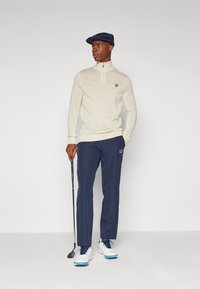 Cream zip-up golf sweater with a small logo, paired with navy golf pants and white shoes, held golf club, wearing a navy cap.
