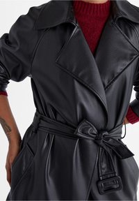 Trench coat nero in ecopelle con collo ampio, vita con cintura, tasche laterali e un indumento interno a maglia con trama visibile sotto.