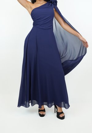 Femme portant une robe longue bleu marine à une épaule avec un détail de cape transparente et des sandales à plateforme à talons hauts noires.