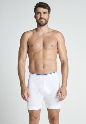 Mand med skæg iført hvide boxer briefs med lange ben står mod en ensfarvet lys baggrund, vendt fremad med afslappet udtryk.