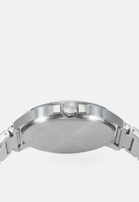 Montre en acier inoxydable avec un boîtier rond, un bracelet argenté, une lunette texturée et une couronne latérale marquée "HUGO". L'arrière du boîtier est lisse.
