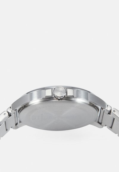 Montre en acier inoxydable avec un boîtier rond, un bracelet argenté, une lunette texturée et une couronne latérale marquée "HUGO". L'arrière du boîtier est lisse.