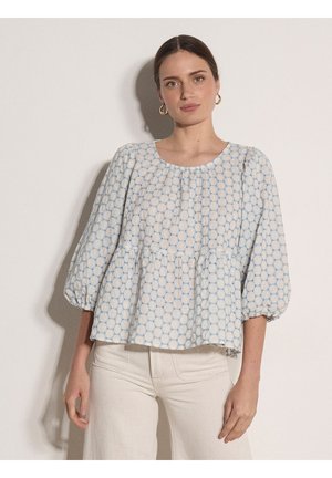 Vrouw met bruin haar in een lichtblauw en wit gebloemd blouse met opgeblazen mouwen, draagt gouden oorringen en witte broek, tegen een effen muur.