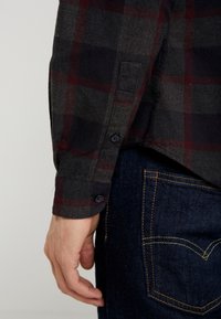 Camicia di flanella a quadri in grigio scuro e bordeaux con polsini abbottonati, abbinata a jeans di denim scuri con cuciture arancioni sulla tasca.