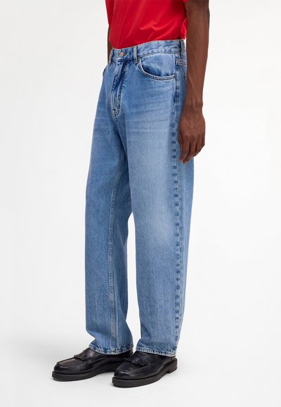Personne portant un jean bleu clair ample, des mocassins noirs et une chemise rouge, debout devant un fond blanc uni.