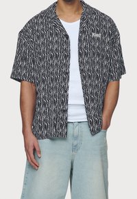 Kortärmad, knappad skjorta gjord av texturerad svart och vit tyg med ett zick-zackmönster. Har en liten logotyp på vänster bröst.