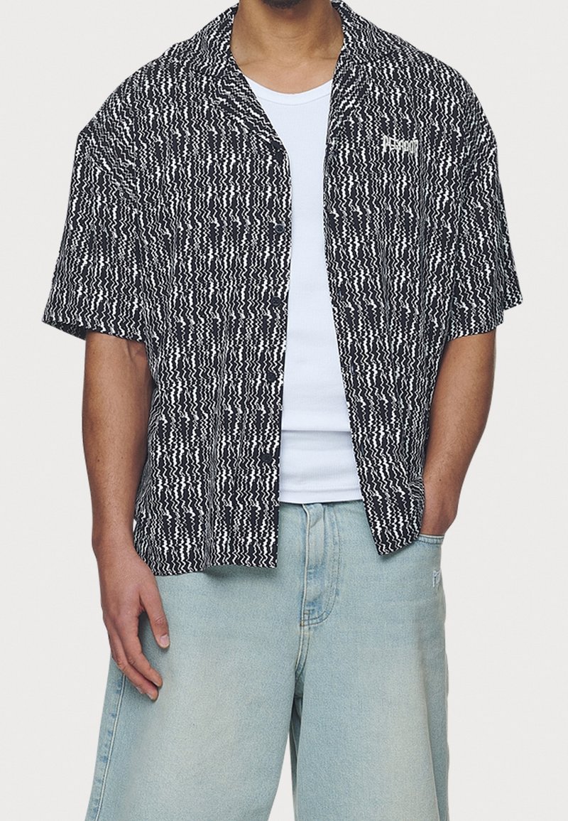 Kortärmad, knappad skjorta gjord av texturerad svart och vit tyg med ett zick-zackmönster. Har en liten logotyp på vänster bröst.