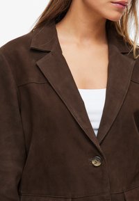 Blazer de camurça castanha com lapelas recortadas, fecho de botão único e detalhes de costura visíveis. Projetado para um ajuste personalizado.
