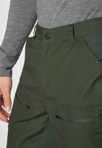 Zielone spodnie cargo o gładkiej fakturze, z dwoma przednimi kieszeniami, zamkowymi detalami oraz zapięciem na guzik w pasie.