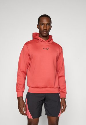 Haut à capuche corail en tissu lisse, avec un logo sur le devant et une capuche à cordon. Associé à un short noir et des accents roses clairs.
