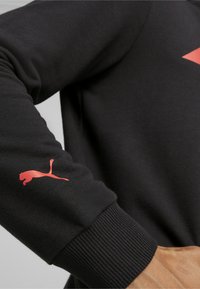 Fekete pulóver bordás mandzsettákkal; a bal ujjon piros Puma logó található; sima textúra és laza dizájn.