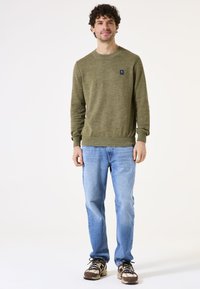 Olivgrüner Pullover mit gerippten Bündchen und Saum, kombiniert mit hellblauen Jeans. Die Sneakers haben ein Muster in Braun, Beige und Schwarz.