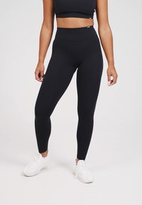 Svarta högmidjade leggings designade för komfort och flexibilitet, tillverkade av mjukt, stretchigt tyg. Kombinerade med vita sportskor.