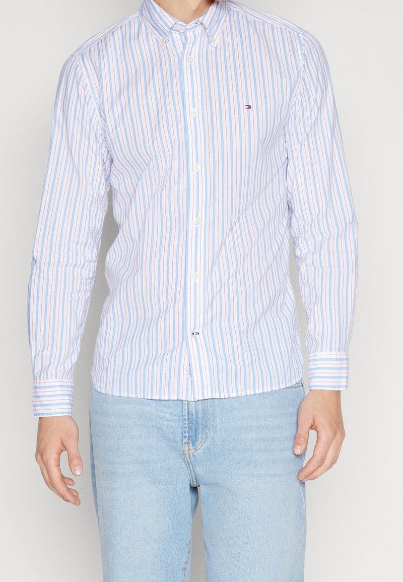 Homme portant une chemise blanche à boutons avec des rayures verticales bleues et rouges et un jean bleu clair, debout devant un fond uni.
